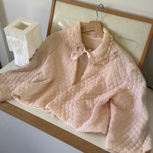 May include: A pale pink quilted jacket featuring a rounded collar with ruffled trim and a satin ribbon tie closure at the neckline. The garment has long sleeves and a textured, diamond-patterned quilted surface throughout the entire fabric.