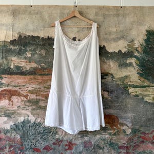 May include: A white cotton slip with lace trim. The slip is hanging on a wooden hanger against a painted backdrop.