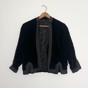 May include: A black velvet jacket with a grey trim, hanging on a wooden hanger. The jacket has a classic design with a collarless neckline and long sleeves. The grey trim is adorned with small, dark embellishments.
