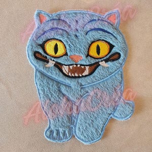 Puede incluir: Parche bordado con un gato azul sonriente con ojos amarillos y una amplia sonrisa. El gato tiene una textura esponjosa y está sentado. El parche es para ropa o accesorios.