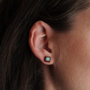 May include: A pair of small square earrings with a turquoise stone set in the center of each earring. The earrings are made of wood and have a dark brown color.