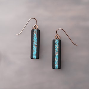 May include: A pair of rectangular earrings with a black base and a turquoise and copper inlay. The earrings have a simple, elegant design with copper-coloured ear wires.