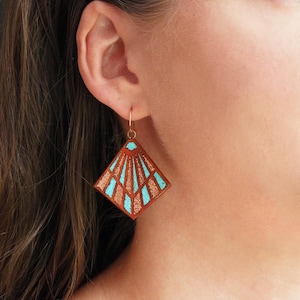 May include: A close-up of a copper-colored earring with a geometric design. The earring features a square shape with a fan-like pattern in turquoise and copper. The earring is hanging from a gold-colored hook.