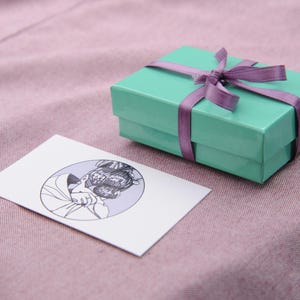 May include: A light green gift box with a purple ribbon and bow. The box is sitting on a pink surface.