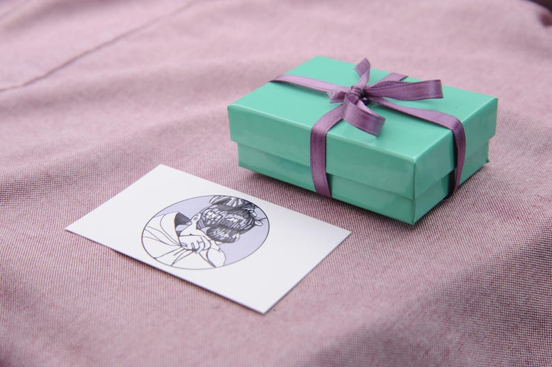 May include: A light green gift box with a purple ribbon and bow. The box is sitting on a pink surface.