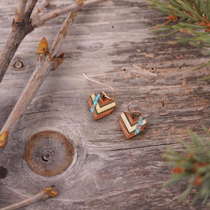 May include: A pair of heart-shaped earrings made of wood with a chevron pattern and turquoise accents. The earrings are on a wooden surface.