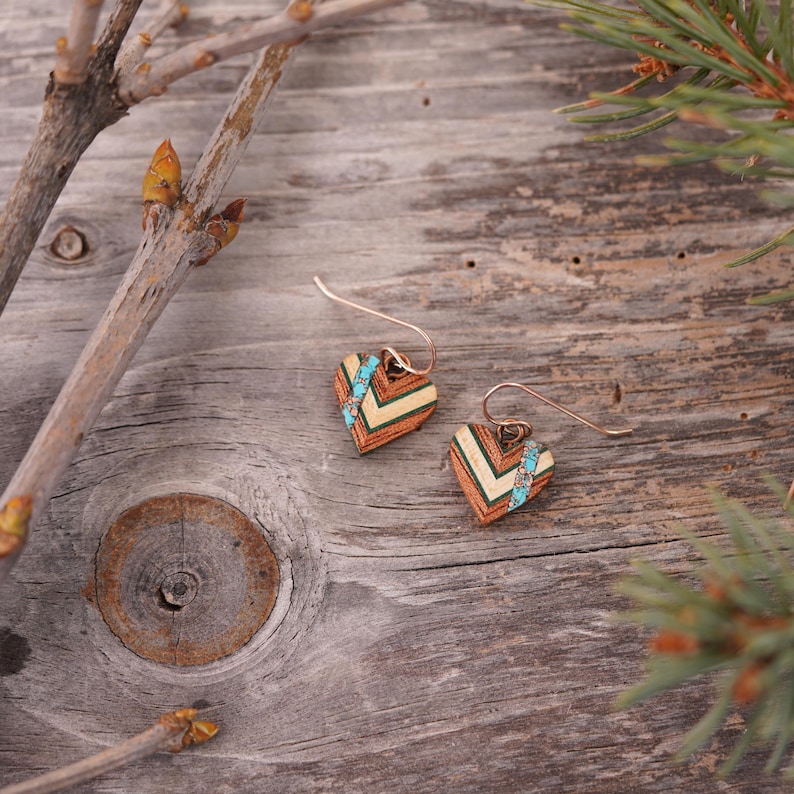 May include: A pair of heart-shaped earrings made of wood with a chevron pattern and turquoise accents. The earrings are on a wooden surface.