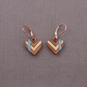 May include: A pair of heart-shaped earrings made of wood with a chevron pattern and a turquoise accent. The earrings have a rose gold finish and are attached to leverback closures.