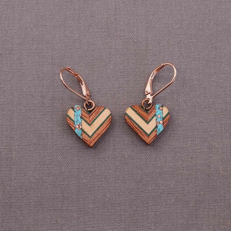 May include: A pair of heart-shaped earrings made of wood with a chevron pattern and a turquoise accent. The earrings have a rose gold finish and are attached to leverback closures.