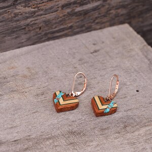 May include: A pair of heart-shaped earrings made of wood with a turquoise inlay. The earrings have a rose gold finish and a leverback closure.
