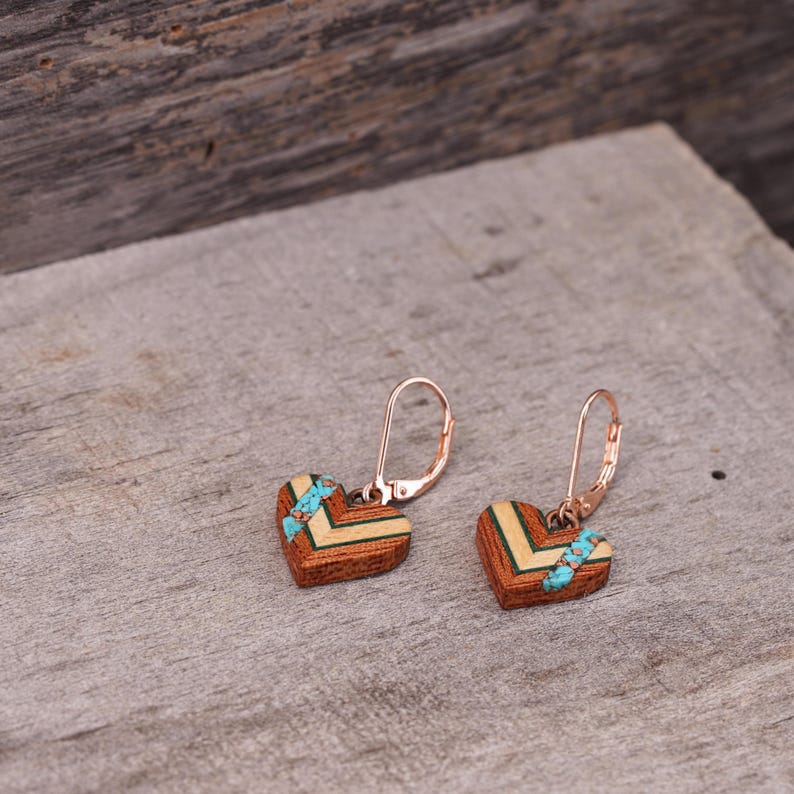 May include: A pair of heart-shaped earrings made of wood with a turquoise inlay. The earrings have a rose gold finish and a leverback closure.
