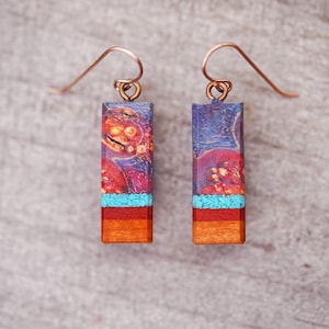 May include: A pair of rectangular earrings with copper-colored hooks. The earrings feature a vibrant design with a mix of colors, including blue, red, orange, and turquoise. The top section has a marbled effect, while the bottom has a layered wood appearance.