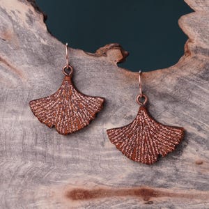 May include: A pair of copper-colored ginkgo leaf earrings with a textured surface. The earrings are hanging from a wooden surface.