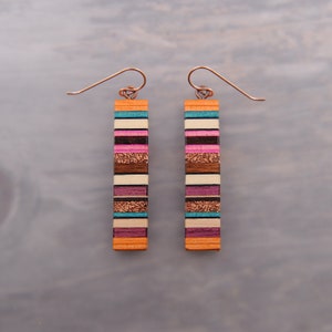 May include: A pair of long, rectangular wooden earrings with a multi-colored striped pattern. The stripes are in shades of pink, orange, brown, white, and blue.