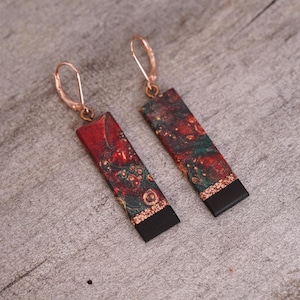 May include: A pair of dangle earrings with a rectangular red, green, and gold marbled design. The earrings have a black accent at the bottom and a rose gold hook.