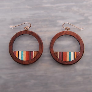 May include: A pair of wooden hoop earrings. The earrings have a dark brown wooden frame with a colorful striped inlay at the bottom. The stripes include shades of blue, red, orange, and brown. The earring hooks are gold-colored.