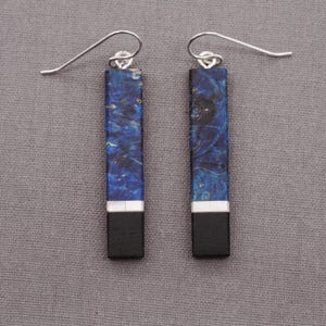 Peut inclure: Une paire de boucles d'oreilles pendantes avec des pièces rectangulaires en bois bleu et noir. Le bois a un motif marbré et une bordure argentée.