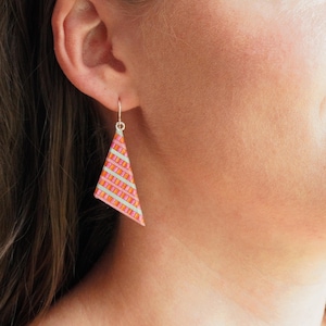 May include: A close-up of a patterned earring. The earring is a triangle shape with a silver hook. The pattern is a mix of pink, orange, and light green stripes. The earring is hanging from a person's ear.