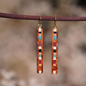May include: A pair of long, rectangular wooden earrings with a geometric pattern of colorful wood inlays. The earrings are suspended from gold-toned hooks.