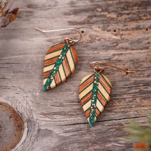 May include: A pair of wooden leaf earrings with a green and brown geometric pattern. The earrings are made of wood and have a copper wire hook.