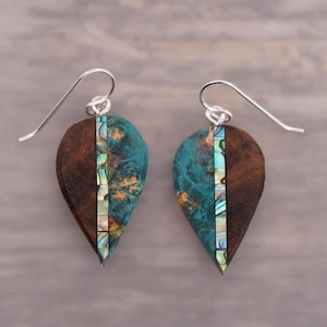 May include: A pair of teardrop-shaped earrings made of wood and abalone shell. The earrings are a dark brown colour with a teal blue and iridescent abalone shell inlay. The earrings are attached to silver hooks.