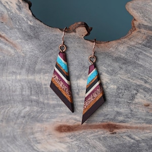 May include: A pair of geometric wood earrings with a layered design. The earrings are made of wood with a dark brown base and stripes of black, white, turquoise, and gold. The earrings are attached to copper hooks.