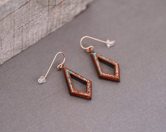May include: A pair of wooden earrings with a diamond shape. The earrings are brown with a turquoise inlay. The earrings are hanging from gold hooks.