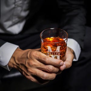 May include: A clear glass with a personalized initial and last name, "M MILLER", is filled with amber-colored liquid. The glass is being held by a person's hands.