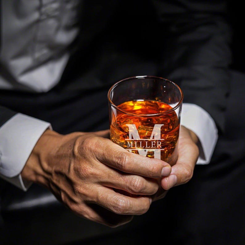 May include: A clear glass with a personalized initial and last name, "M MILLER", is filled with amber-colored liquid. The glass is being held by a person's hands.