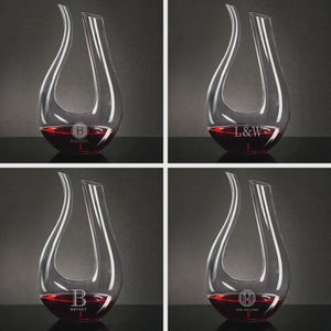 May include: Four clear glass wine decanters, each filled with red wine. Each decanter has a unique monogram or name etched on the base. The decanters have a teardrop shape with a wide base and a narrow opening.