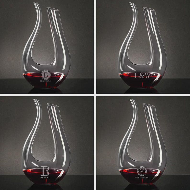 May include: Four clear glass wine decanters, each filled with red wine. Each decanter has a unique monogram or name etched on the base. The decanters have a teardrop shape with a wide base and a narrow opening.