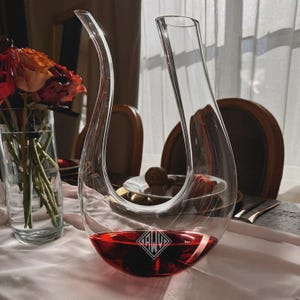 May include: A clear glass wine decanter with a curved spout and a wide base, filled with red wine. The decanter has a diamond-shaped logo on the base. The decanter is on a white tablecloth, with flowers in a vase in the background.