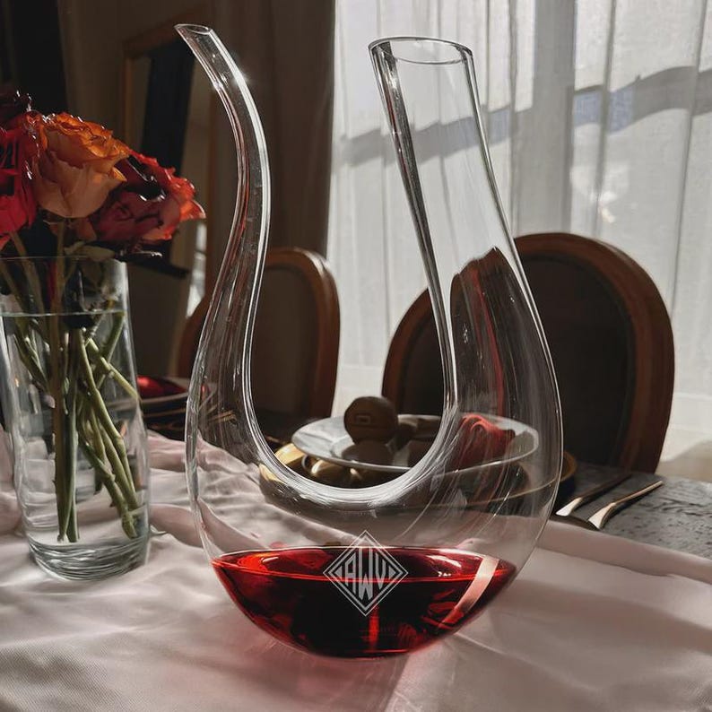 May include: A clear glass wine decanter with a curved spout and a wide base, filled with red wine. The decanter has a diamond-shaped logo on the base. The decanter is on a white tablecloth, with flowers in a vase in the background.