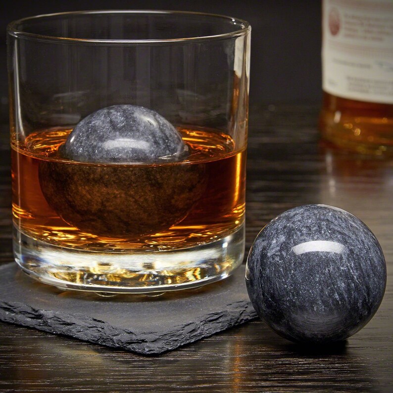 May include: Two gray marble whiskey stones in a glass of amber liquid. One stone is in the glass, the other is on a black slate coaster.