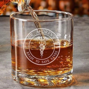 May include: A clear glass rocks glass with a personalized inscription that reads "Dr. Andrews Ready to Save Lives" with a medical symbol in the center.