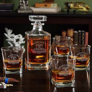 May include: A set of five clear glass decanter and glasses with a personalized design. The decanter and glasses are engraved with the text "Maddison's Distilled Spirits" and a monogram. The glasses are square shaped and the decanter is square with a round top.