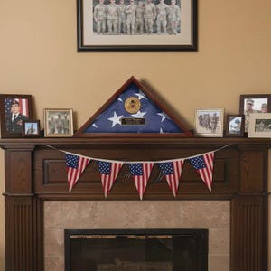 May include: A wooden mantelpiece decorated with framed photos and a triangular flag display case. A string of American flag pennants hangs below. Above, a framed group photo of soldiers is displayed.