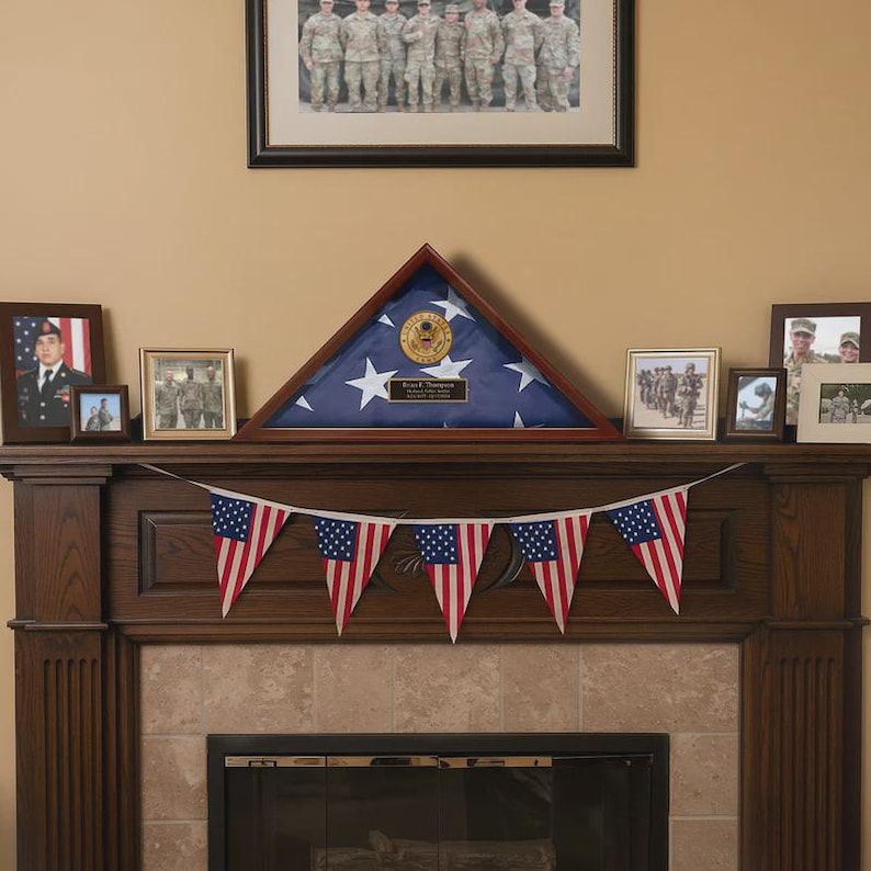 May include: A wooden mantelpiece decorated with framed photos and a triangular flag display case. A string of American flag pennants hangs below. Above, a framed group photo of soldiers is displayed.