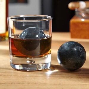 May include: A clear glass with a dark brown liquid and a large black ice sphere inside. A smaller black ice sphere sits on a wooden surface next to the glass.