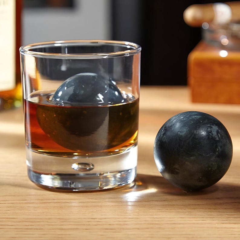 May include: A clear glass with a dark brown liquid and a large black ice sphere inside. A smaller black ice sphere sits on a wooden surface next to the glass.