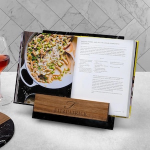 May include: A cookbook holder with a wooden base and a black support. The cookbook is open to a recipe with a photo of pasta. A wooden nameplate with the name "Fitzpatrick" is in front of the book.