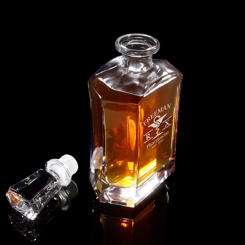May include: A clear glass decanter with a stopper, filled with amber liquid. The decanter is engraved with the text "FREEMAN R2X Pharmacist Est. 2021" and a stylized Rx symbol.