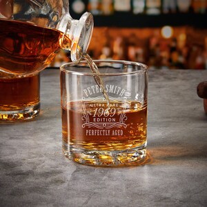 May include: A clear glass whiskey tumbler with the text "Peter Smith Ultra Rare 1969 Edition Perfectly Aged" etched on the side. The glass is filled with amber liquid and a decanter is pouring more liquid into the glass.