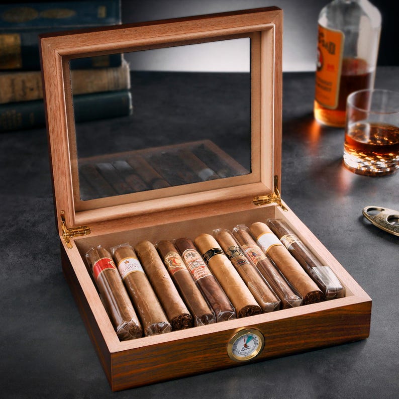May include: A wooden cigar box with a glass top and a hygrometer. The box is filled with ten cigars, each wrapped in cellophane. The cigars are arranged in two rows of five.