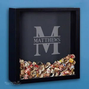 May include: A black shadow box with a gray background and the name "MATTHEWS" in a split monogram design. The box is filled with cigar bands.