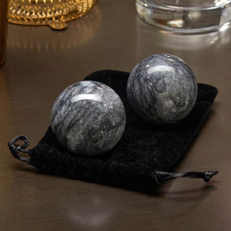 May include: Two gray marble massage balls with black veining, resting on a black velvet pouch. The balls are smooth and round, and the pouch is soft and plush.