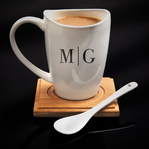 K&ouml;nnte beinhalten: Wei&szlig;e Keramiktasse mit schwarzer Aufschrift "M|G" und einer braunen Fl&uuml;ssigkeit im Inneren. Die Tasse steht auf einem Untersetzer aus Holz mit einem wei&szlig;en Keramikl&ouml;ffel.