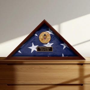 May include: A triangular wooden flag display case. Inside, a folded American flag with white stars on a blue field is visible. A gold emblem and a nameplate with the text "Brian F. Thompson" are also present. The case is displayed on a wooden surface.