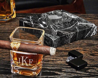 May include: A black marble ashtray with a square shape and a round depression in the center. A clear glass with a monogrammed "JKF" sits on a wooden surface with a brown cigar resting on the rim of the glass. A black lighter is also on the surface.