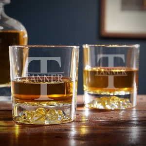 May include: Two clear glass whiskey tumblers with a textured bottom. The glasses are filled with amber liquid and have the letter "T" and the name "Tanner" etched on the front.